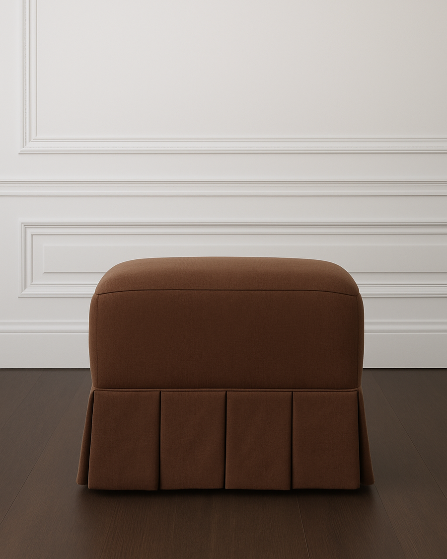 Sloane Ottoman in Espresso Linen with pleated skirt on a dark wood floor against a white panelled wall.