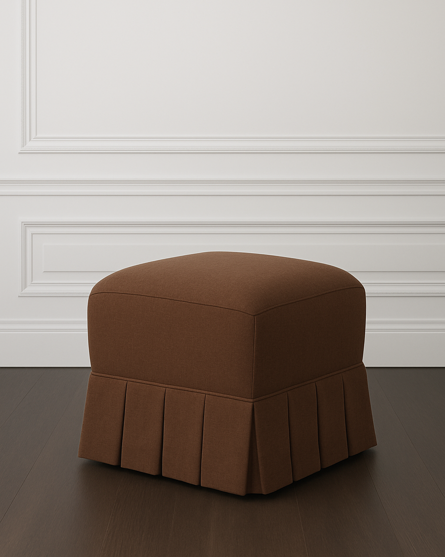 Sloane Ottoman in Espresso Linen with pleated skirt on a dark wood floor against a white panelled wall.